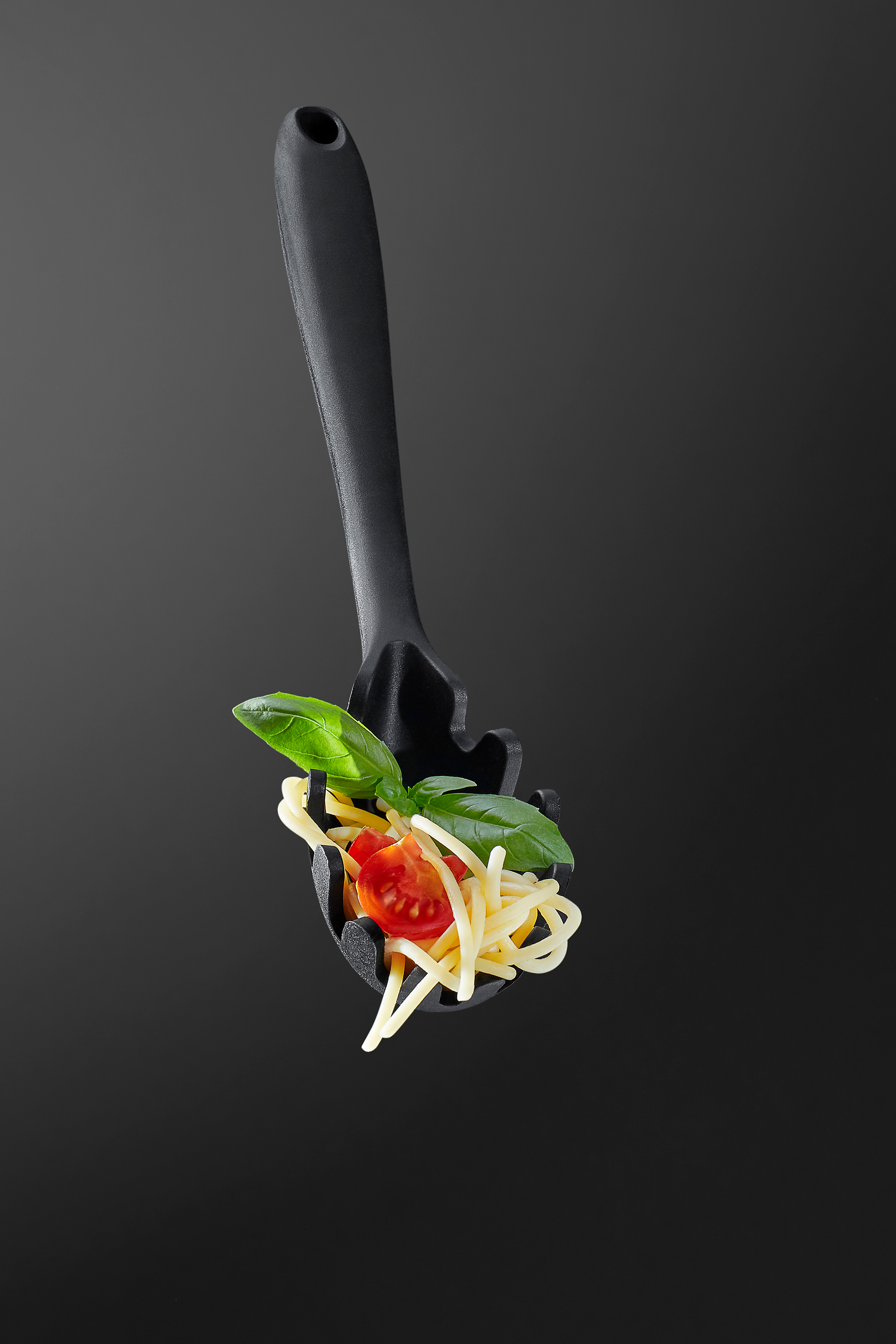 photo of a spaghetti spoon hanging in the air filled with pasta, tomato and basil leafs shot on a black background